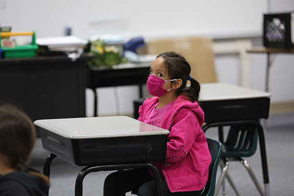 little girl in a pink jacket and mask sitting at her desk in her classroom
